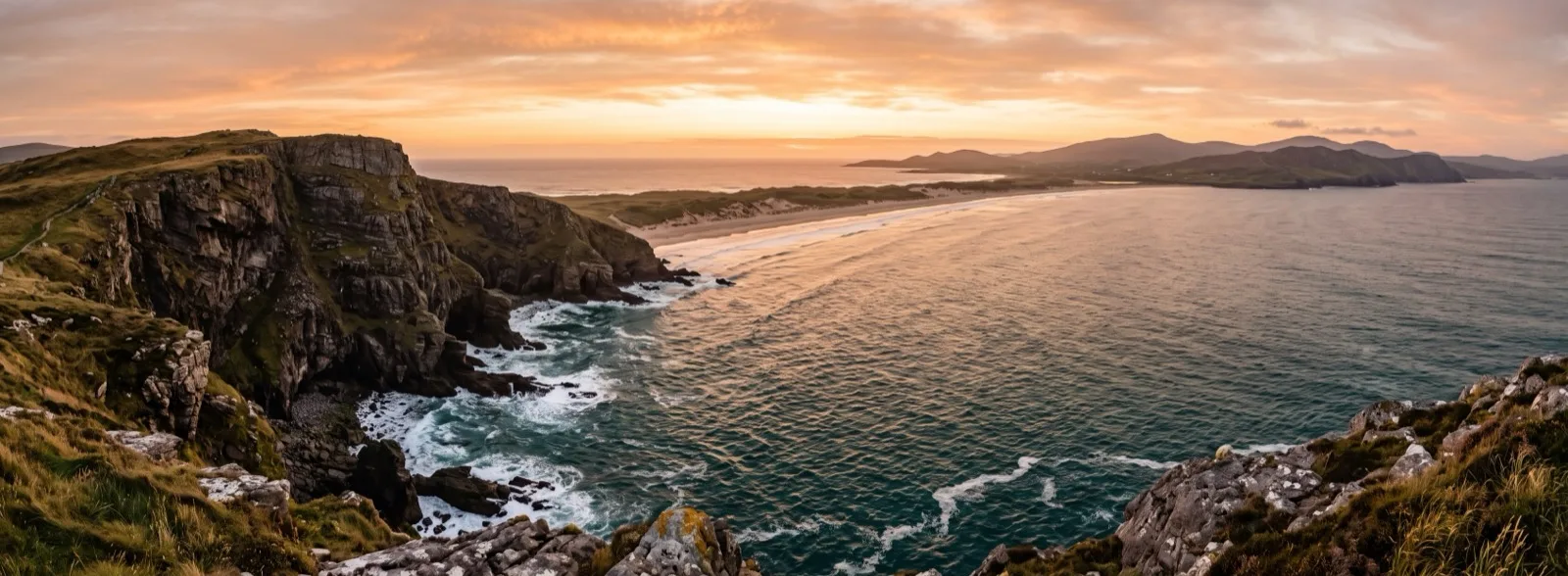 A wide coastal panorama: cliffs on the left, a curving bay in the centre and distant hills on the right, joined into one continuous view